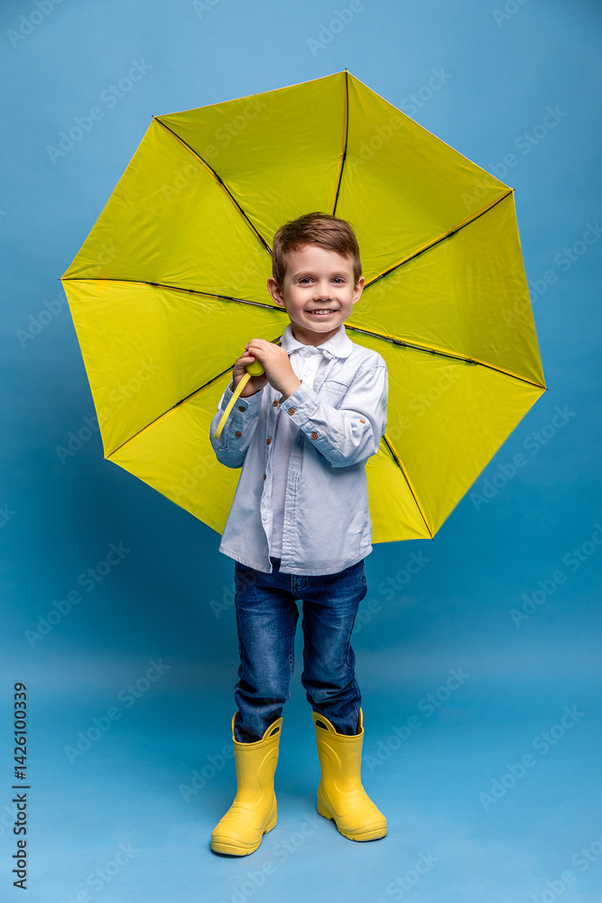 Happy funny smiling child, little boy, with yellow umbrella posing on blue background. Kid is wearing yellow rubber boots. Rainy spring or autumn weather. Clothes for the rain. Copy space