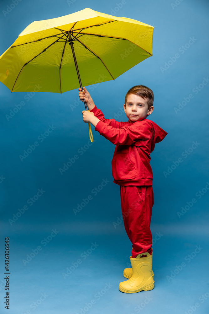 Happy funny smiling child, little boy in red sport suit with yellow umbrella posing on blue background. Kid is wearing yellow rubber boots. Rainy spring or autumn weather. Copy space