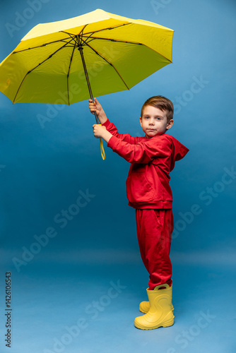 Obraz na płótnie Happy funny smiling child, little boy in red sport suit with yellow umbrella posing on blue background. Kid is wearing yellow rubber boots. Rainy spring or autumn weather. Copy space w salonie