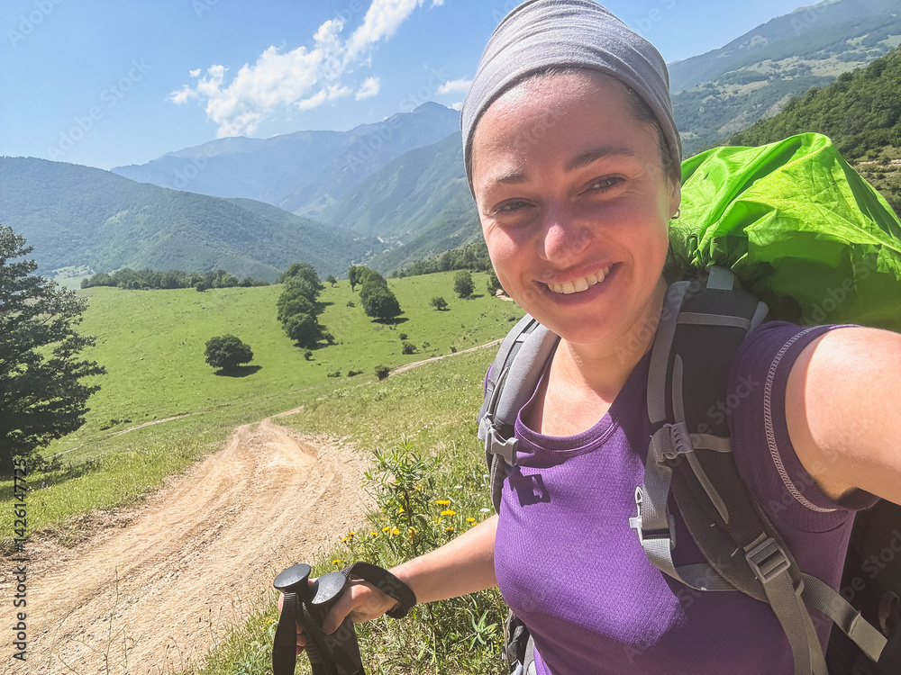 Obraz premium Hiking tourism adventure. Backpacker hiker woman taking selfie picture. Hiker girl lady tourist with backpack walking near mountain. Young happy woman enjoy hike trekking tourism active vacation