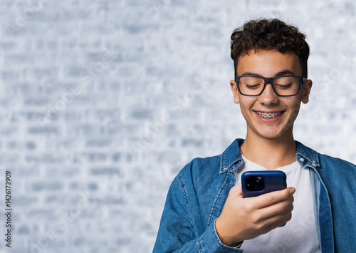 Smiling black curly hair funny young man, guy, boy wear braces hold hand typing text, looking reading gazing at cell phone mobile smartphone cellphone, stand against white bricks loft wall background