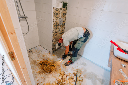 Worker removing rotted subfloor in a bathroom during renovation. Visible damage from water exposure. Tools and debris scattered around..