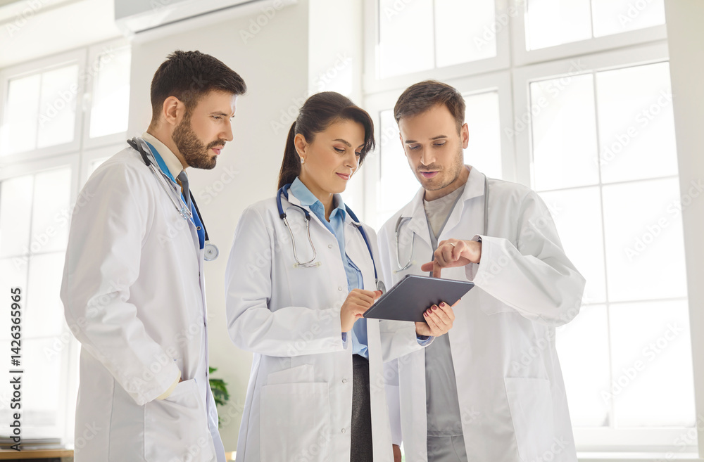Three professional doctors gathered in medical office looking at tablet screen and discussing details. Focused clinic staff in lab coats reviewing test results and collaborating on treatment decisions