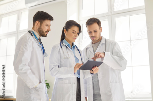 Obraz na płótnie Three professional doctors gathered in medical office looking at tablet screen and discussing details. Focused clinic staff in lab coats reviewing test results and collaborating on treatment decisions w salonie