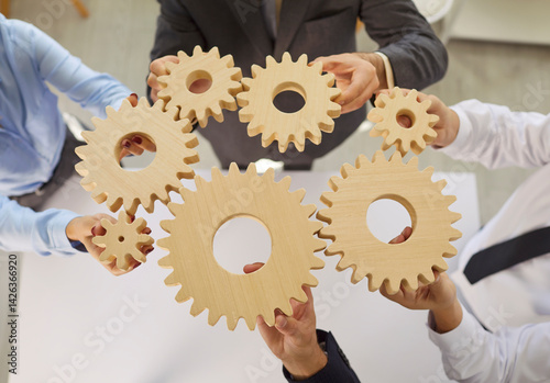 In office environment, several business people hold various wooden gears together over meeting table. Colleagues demonstrate importance of teamwork and strategic planning in business operations.
