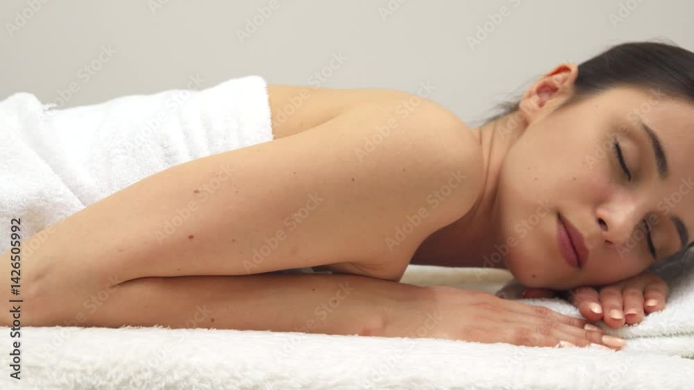 A beautiful brunette woman lies peacefully on her stomach on a plush massage table, wrapped in a soft white towel. Her serene expression and closed eyes evoke a sense of tranquility, inviting viewers A beautiful brunette woman lies peacefully on her stomach on a plush massage table, wrapped in a soft white towel. Her serene expression and closed eyes evoke a sense of tranquility, inviting viewers