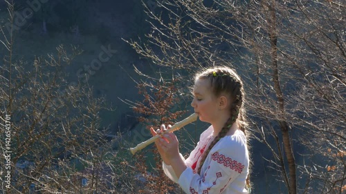 A little girl in a vyshyvanka, a Ukrainian embroidered shirt, playing the flute outdoors. A child learning recorder or block flute, a woodwind musical instrument. Hobby, flute lesson, musical training