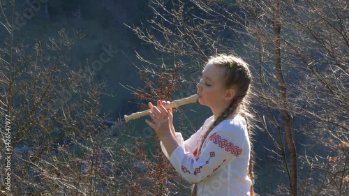 A little girl in a vyshyvanka, a Ukrainian embroidered shirt, playing the flute outdoors. A child learning recorder or block flute, a woodwind musical instrument. Hobby, flute lesson, musical training