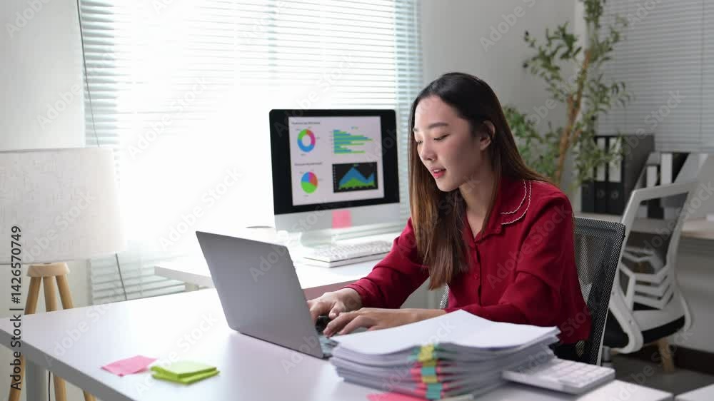 A woman is sitting at a desk with a laptop and a stack of papers. She is focused on her work and she is in a serious mood A woman is sitting at a desk with a laptop and a stack of papers. She is focused on her work and she is in a serious mood
