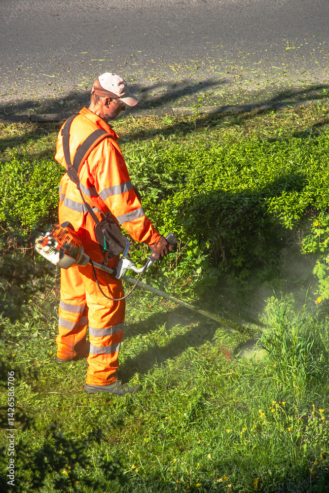 Man in orange uniform and protective glasses mowing green grass with a lawnmower on a sunny day. Bright green lawn and natural outdoor background.