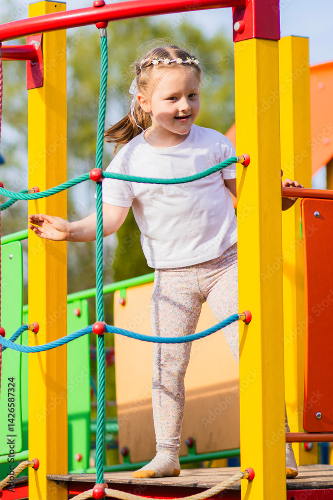 Happy blonde girl in a white T-shirt with a floral headband smiling joyfully on a vibrant, colorful playground. The background is full of playful red, yellow, green, and blue elements.