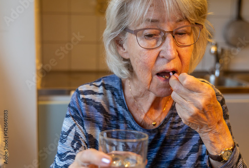 Papier peint An elderly, gray-haired woman takes her medication, holding a glass of water in one hand and a pill in the other