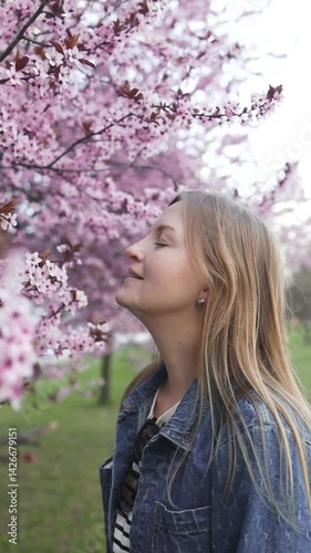 A smiling woman stands against a backdrop of blooming cherry blossoms in spring. The vibrant pink flowers create a serene, picturesque atmosphere as she enjoys the beautiful season outdoor