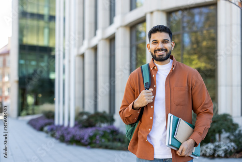Obraz na płótnie A smiling student poses outside a modern building, holding books and a backpack, ready for classes at a university campus. The scene depicts a beautiful sunny day. w salonie