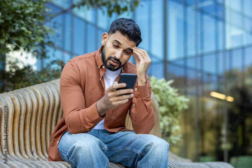 A concerned man looks at his phone while sitting outside on a bench. The scene shows a modern building in the background.