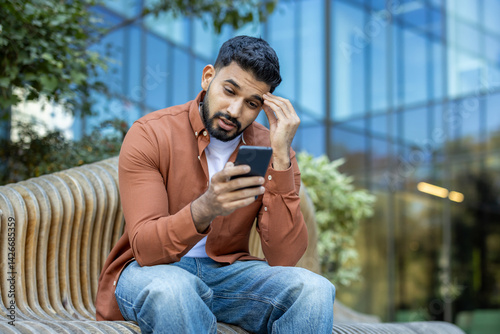 Obraz na płótnie A concerned man looks at his phone while sitting outside on a bench. The scene shows a modern building in the background. w salonie