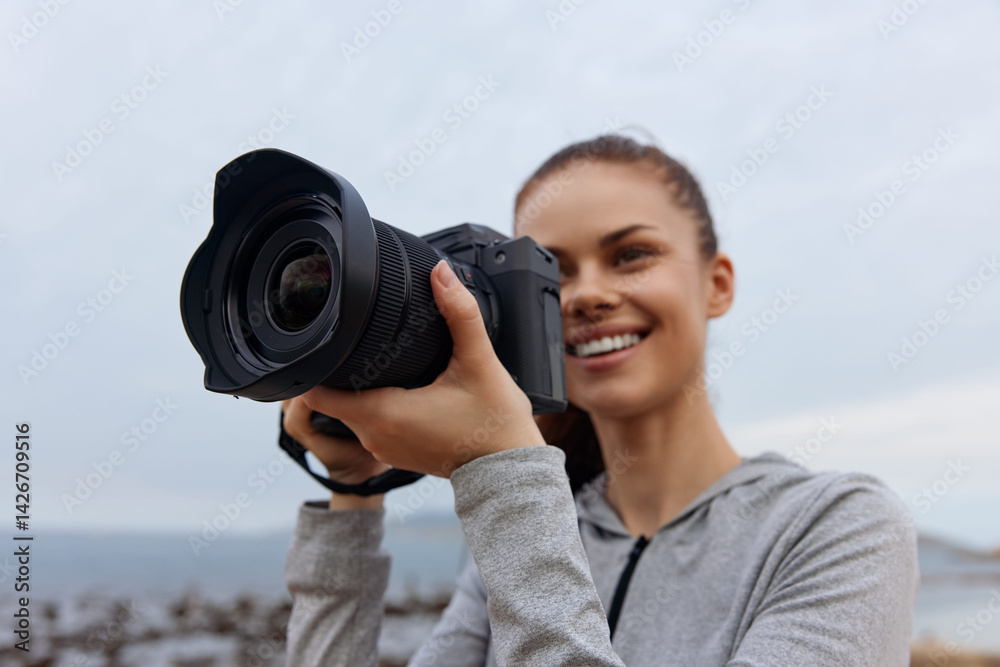 Obraz premium Young woman smiling while holding a professional camera by the beach, showcasing a passion for photography and a joyful connection with nature