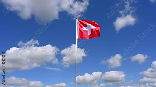 The Swiss flag is hoisted and flutters in the wind against a blue sky with white clouds, 4K, real time
