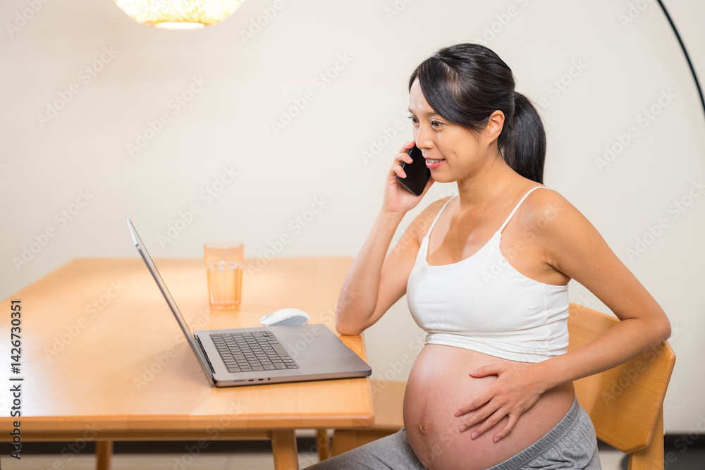 Pregnant woman working on laptop and talking on phone at home