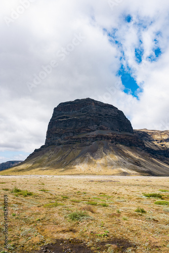 Tall, stratified volcanic rock formation in Icelandic wilderness, with mossy tundra and moody skies above.