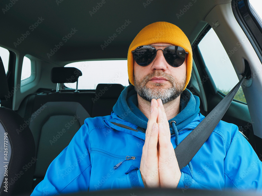 Man driver in blue jacket and yellow hat wearing dark aviator glasses. Travel and transport. Male model with grey beard, hands in prey gesture. Road safety theme.