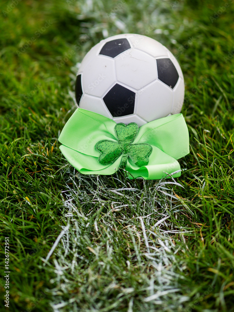 Green bow with Irish shamrock by a classic football or soccer ball on a green grass training field. Goal post in the background. Supporting national team of Ireland concept.