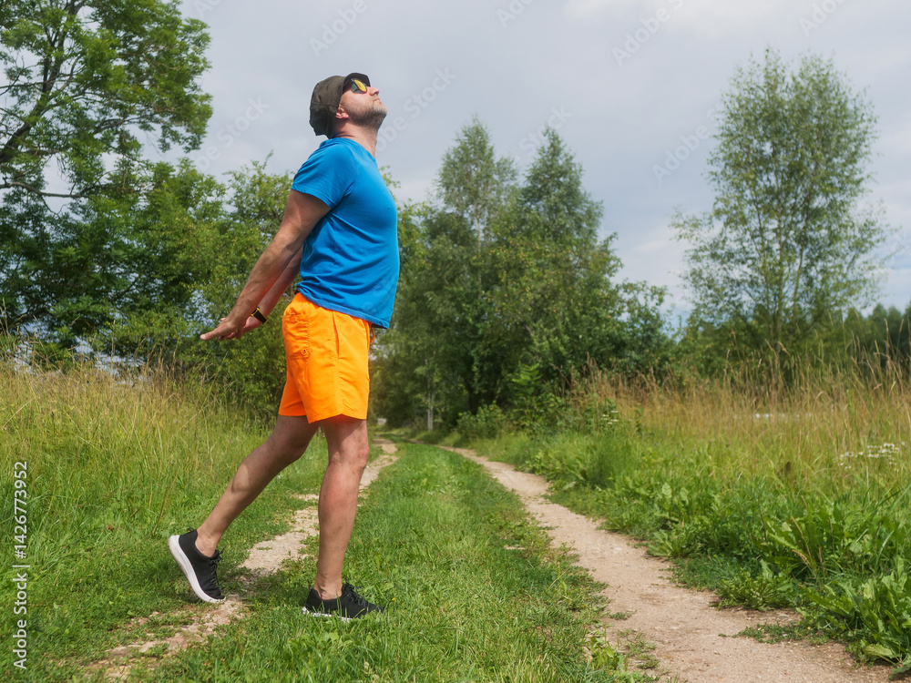 A man in a blue shirt and orange shorts stretching his back on a country road. The man is wearing a hat and he is enjoying the outdoors. Male in his 40s with grey beard. Sport and fitness outdoor.