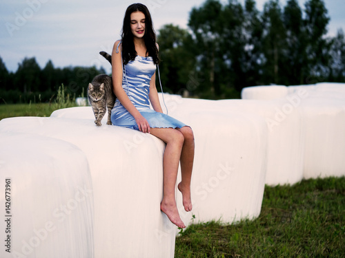 Young teenager girl in high fashion dress on a white plastic bale with hey and her pet cat. Glamour photoshoot in a country side area at dusk. Model is slim with long hair. Pretty girl outdoors.
