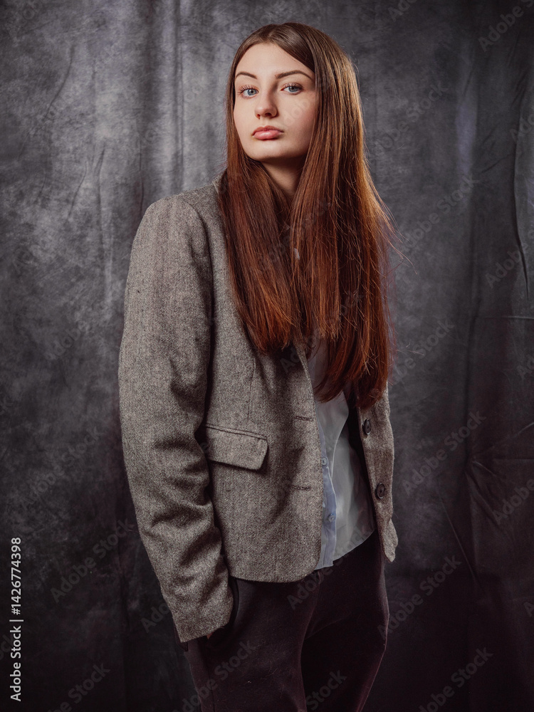 A young business woman is wearing a gray jacket and black pants. The model has long brown hair is elegant and is standing in front of a gray color background. Lady boss concept. Classic portrait.