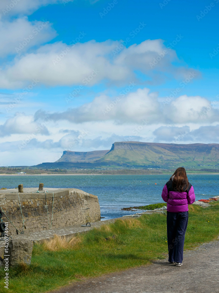 Travel in Ireland. Girl tourist looking at stunning Benbulbin flat top mountain in county Sligo, Ireland. Warm day with rich blue cloudy sky. Amazing Irish nature scenery. Explore nature concept.