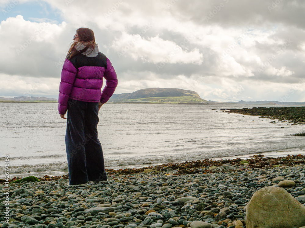 Travel in Ireland. Young girl standing on rough stone beach and looking at ocean and Knocknarea mountain in the background. Stunning Irish nature. Cloudy sky. Famous landmark in county Sligo