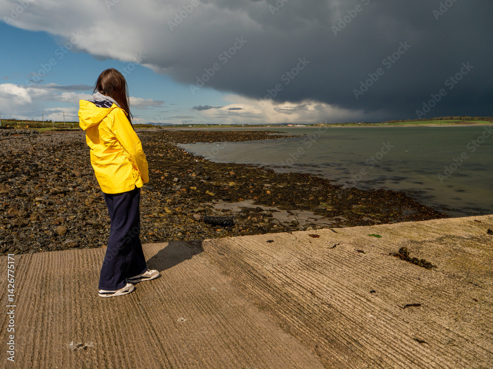 Teenager girl in yellow travel jacket standing on a pier. Huge dark storm cloud in the background. Travel in harsh weather conditions with sun and rain. County Sligo, Ireland.