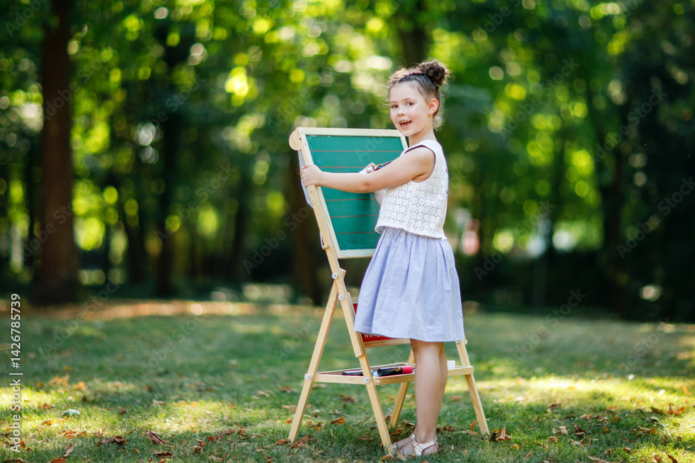 Happy little kid girl standing by big chalk desk Preschool or schoolkid on first day of elementary class. Back to school concept. Healthy child writing and painting on desk outdoors. Copyspace on desk