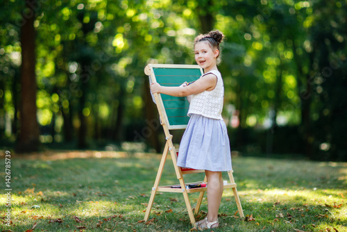 Obraz na płótnie Happy little kid girl standing by big chalk desk Preschool or schoolkid on first day of elementary class. Back to school concept. Healthy child writing and painting on desk outdoors. Copyspace on desk w salonie