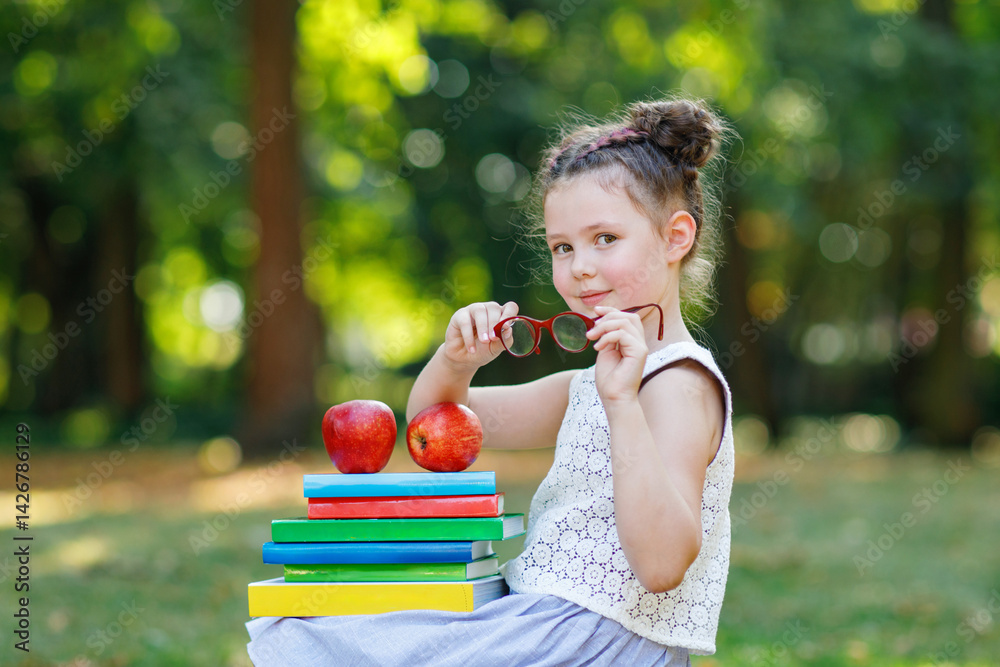 Happy adorable little kid girl reading book and holding different colorful books, apple and glasses on first day to school or nursery. Back to school concept. Healthy child of elementary class.
