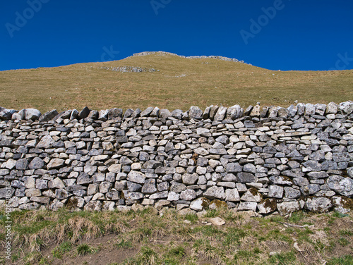 Dry stone wall in the Yorkshire Dales, constructed from irregularly shaped stones stacked without mortar, with a grassy hill rising gently towards the horizon under a clear blue sky.