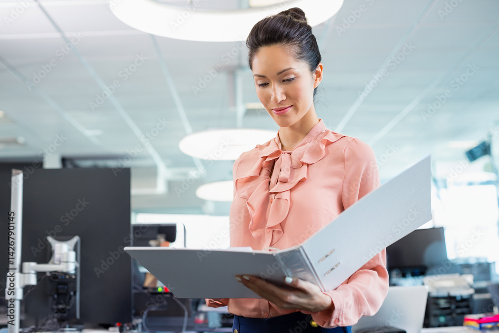 Young Asian woman reading ring binder in open plan office with computer workstation, copy space