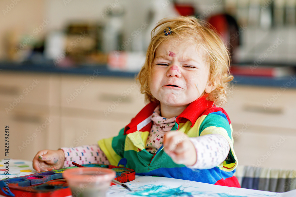 Sad crying baby girl learning painting with water colors. Frustrated Little toddler child drawing at home, using colorful brushes. Angry child in hysteric crisis.