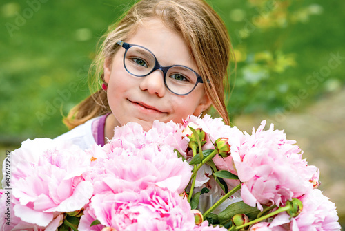 Obraz na płótnie portrait of a happy cute little caucasian seven year old kid girl, holds in hands and smell and enjoying a bouquet of pink peony flowers in full bloom on the background of nature w salonie