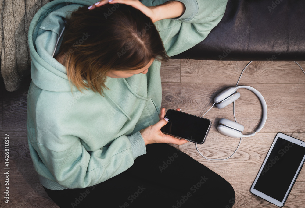 Beautiful young woman in a hoodie is using a smart phone while sitting on the floor near the couch and relaxing, top view