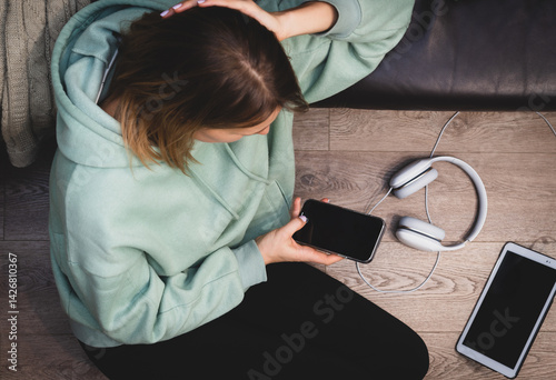 Obraz na płótnie Beautiful young woman in a hoodie is using a smart phone while sitting on the floor near the couch and relaxing, top view w salonie