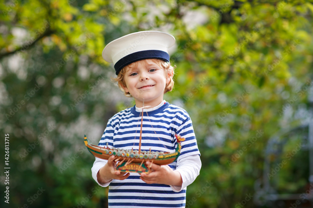 Happy little kid boy in sailor capitain hat and uniform playing with sailor boat ship. Smiling preschool child dreaming and having fun. Education, profession, dream concept