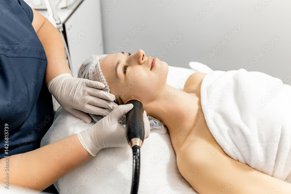 Obraz premium Young woman undergoing radio frequency facelift procedure. Concept of advertising facial skin care, anti-aging facial rejuvenation. Radio wave facelift in cosmetology clinic. RF lifting