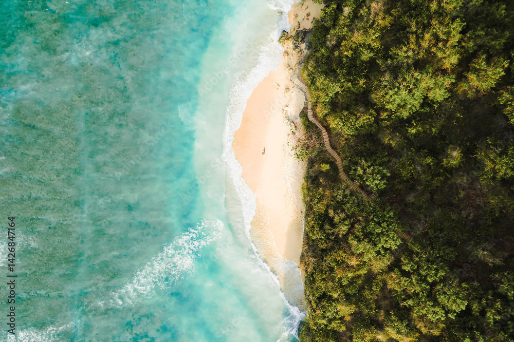 Drone shot of a secluded beach in Bali, with crystal-clear water, crashing waves, and dense tropical vegetation along the rugged coastline.