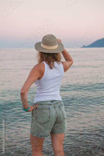 Woman in hat stands barefoot in ocean at sunset, gazing into distance