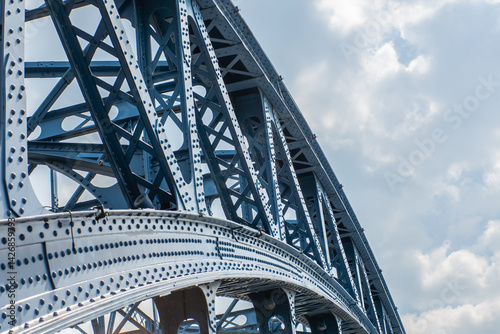 Close-up view of a steel truss bridge with detailed riveted construction, showcasing the geometric precision and strength of modern industrial architecture against a cloudy sky