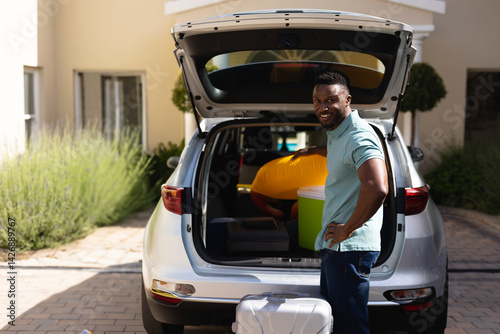 African American man standing by silver SUV trunk in driveway, with cooler and suitcase, copy space