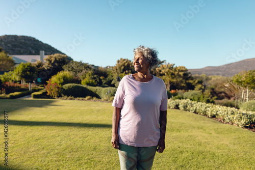 Senior woman standing and looking left in residential garden, enjoying sunshine and lush greenery