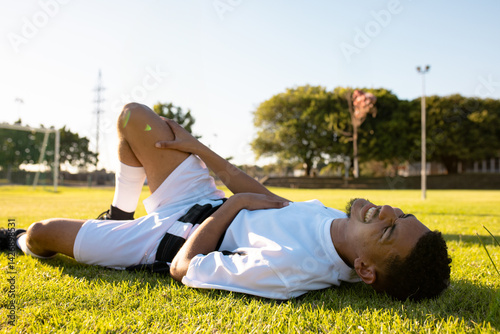 Male soccer player lying on soccer pitch grass near goal frame clutching knee in white jersey
