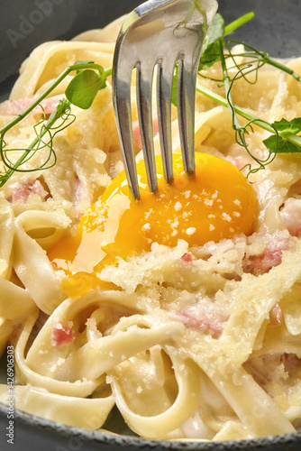 Fork piercing egg yolk in creamy carbonara pasta